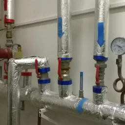 Izolowane rury z zaworami i manometrem, zabezpieczone folią aluminiową, w pomieszczeniu z białymi ścianami. Elementy instalacji grzewczej.