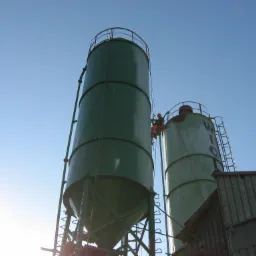 Pracownik zabezpieczony linami, w trakcie pracy na wysokości na tle dwóch silosów w odcieniach zieleni i błękitnego nieba.