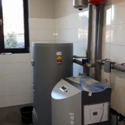 Szara pompa ciepła SmartHeat z zbiornikiem w pomieszczeniu z białymi kafelkami, widok na okno i instalację grzewczą.