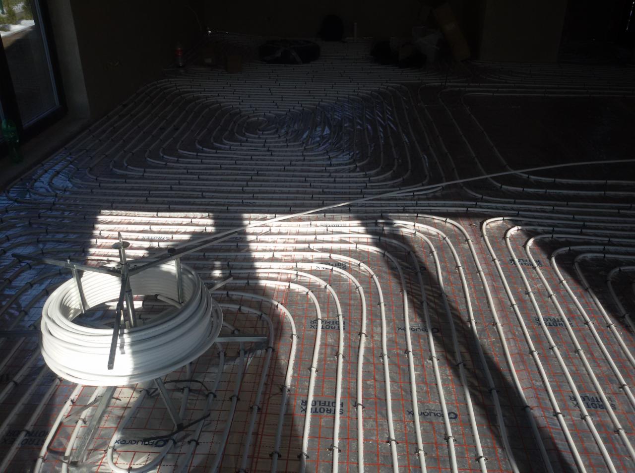 Białe rury ogrzewania podłogowego ułożone na czerwonej macie z siatką, częściowo oświetlone promieniami słońca wpadającymi przez okno.