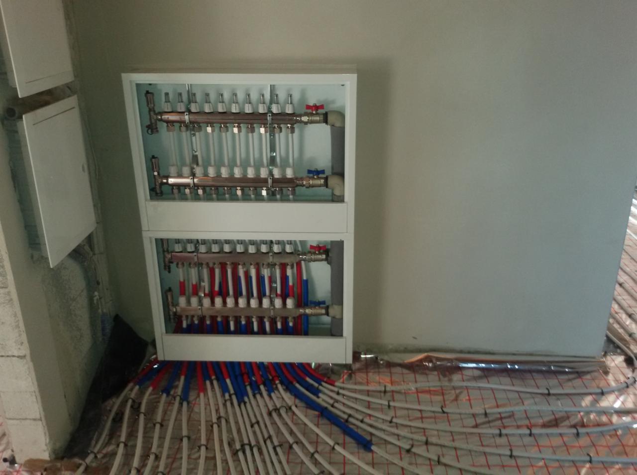 Rozdzielacz ogrzewania podłogowego z rurami PEX w kolorach czerwonym i niebieskim, ułożonymi na siatce grzewczej. Biała szafka z zaworami na ścianie.