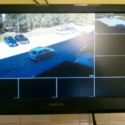 Monitor z podglądem z kamer monitoringu na zewnątrz budynku, widoczne samochody i fragment otoczenia.
