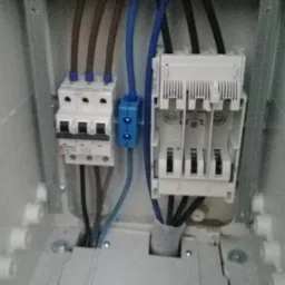 Wnętrze skrzynki elektrycznej z zamontowanymi bezpiecznikami i przewodami w kolorach brązowym i niebieskim, widoczne śruby mocujące i naklejka z kodem kreskowym na dolnej pokrywie.