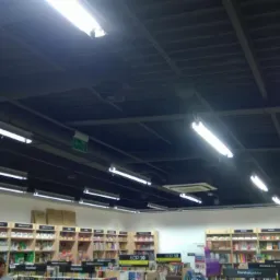 Równomiernie rozmieszczone, podwieszane lampy fluorescencyjne oświetlają sufit w księgarni, widoczne regały z książkami poniżej.