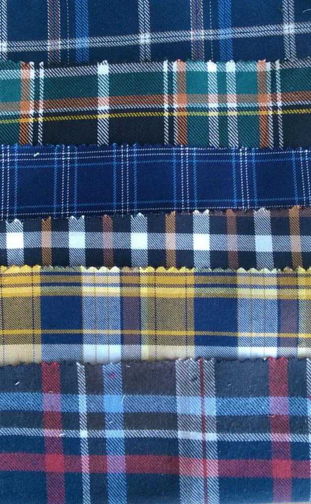 Próbki tkanin koszulowych w kratę o różnych wzorach i kolorach, ułożone warstwami, z widocznymi postrzępionymi brzegami.