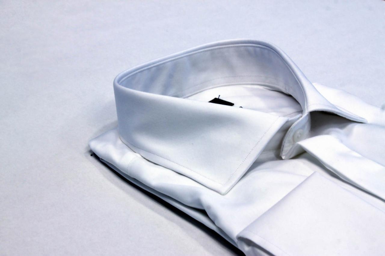 Biała, elegancka koszula męska z mankietami i wykrochmalonym kołnierzykiem, ułożona na jasnym tle, widok z bliska.