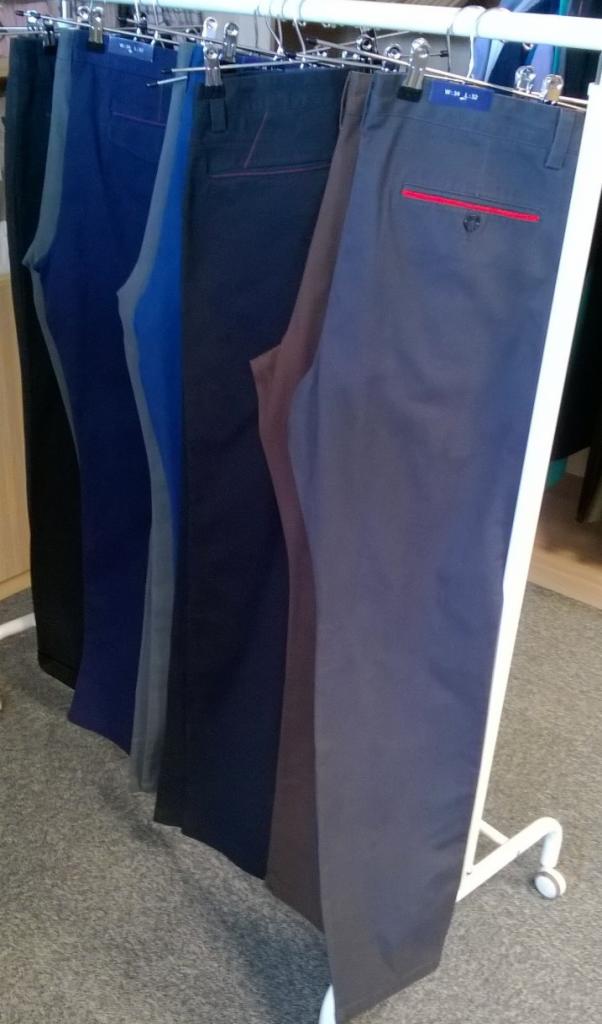 Rząd spodni młodzieżowych w różnych odcieniach niebieskiego, szarego i brązowego, wiszących na metalowym wieszaku z klamerkami, z detalami w postaci czerwonych akcentów na tylnych kieszeniach.