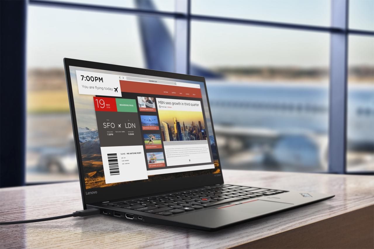 Czarny laptop Lenovo z wyświetlonym biletem lotniczym i artykułem biznesowym, ustawiony na drewnianym biurku na tle okna lotniska z widocznym samolotem.