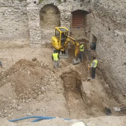 Prace ziemne przy fundamentach starego, kamiennego budynku z użyciem mini koparki Komatsu i łopat. Widoczni pracownicy w kamizelkach odblaskowych.