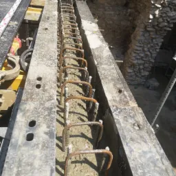 Zbrojenie w szalunku zalane betonem, widoczne metalowe pręty zbrojeniowe i betonowa masa, w tle kamienny mur z łukiem.