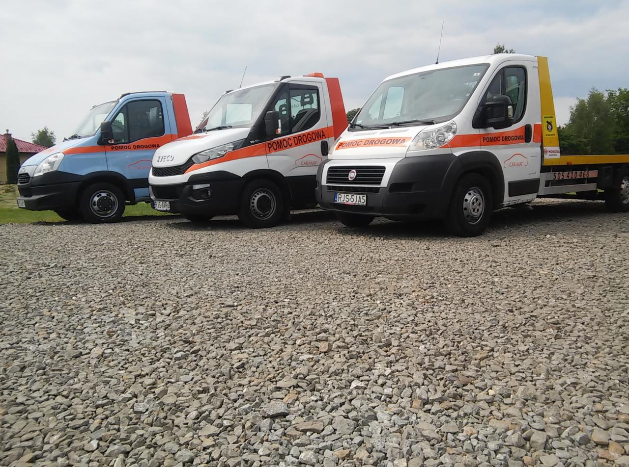 Trzy lawety pomocy drogowej różnych modeli (Iveco i Fiat) stojące na żwirowym parkingu, z widocznymi numerami telefonów i oznaczeniami firmy CAR-MAT.