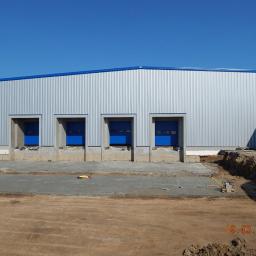 Hale Korman Sp. z o.o. Sp.k. - Nowa hala z blachy trapezowej z trzema bramami w kolorze niebieskim, widok z zewnątrz w słoneczny dzień. Na pierwszym planie teren budowy z narzędziami.