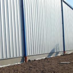 Hale Korman Sp. z o.o. Sp.k. - Długa, srebrna hala przemysłowa z niebieskimi akcentami, widoczna z niskiej perspektywy, z ziemią na pierwszym planie. Widoczne rury odprowadzające wodę.