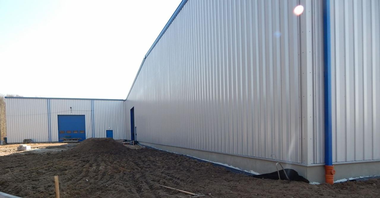 Nowoczesna hala w srebrnej blasze trapezowej z niebieskimi akcentami. W tle widoczne drzewa, a na pierwszym planie ziemia i narzędzia budowlane.