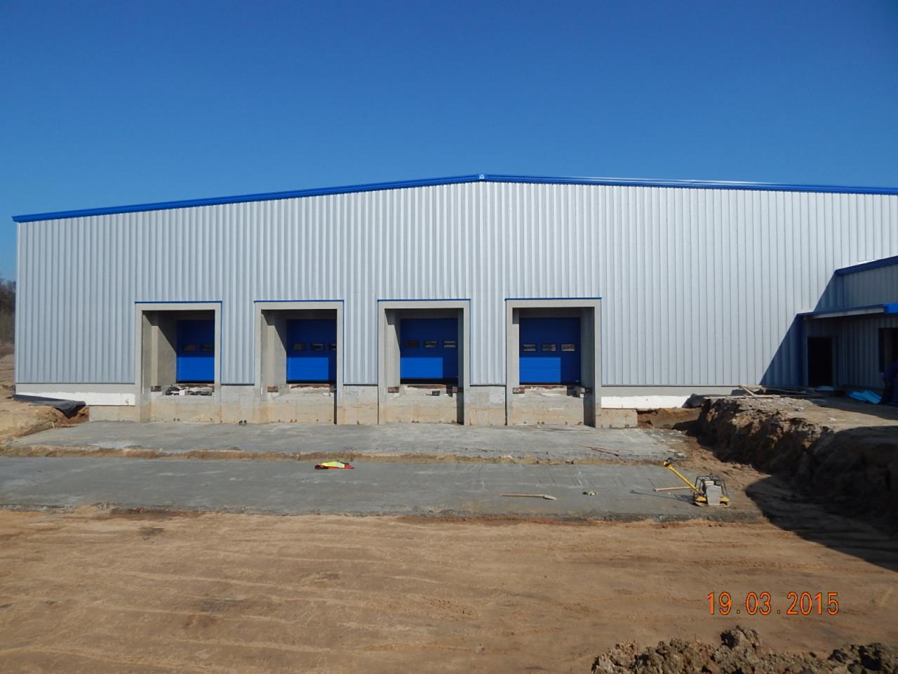 Nowa hala z blachy trapezowej z trzema bramami w kolorze niebieskim, widok z zewnątrz w słoneczny dzień. Na pierwszym planie teren budowy z narzędziami.