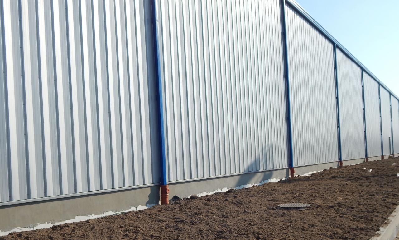 Długa, srebrna hala przemysłowa z niebieskimi akcentami, widoczna z niskiej perspektywy, z ziemią na pierwszym planie. Widoczne rury odprowadzające wodę.