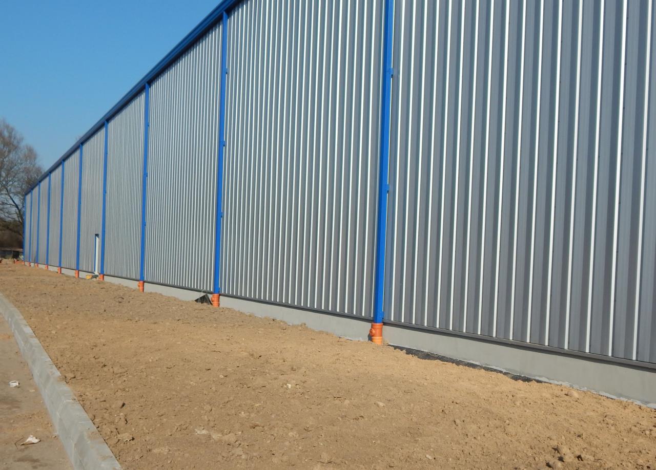 Długa hala z blachy trapezowej, niebieskie słupy wzmacniające, widoczne elementy instalacji u dołu ściany, ziemia wokół. Ujęcie z dołu, błękitne niebo.