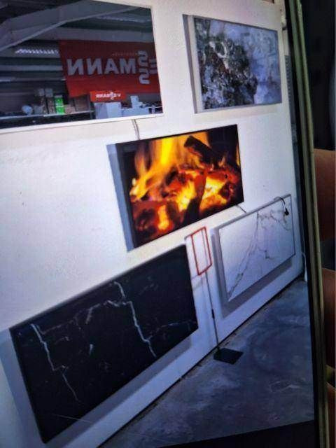 Ekspozycja mat grzewczych z różnymi wzorami (imitacja marmuru, ogień) na ścianie w sklepie z widocznym logo producenta w tle.