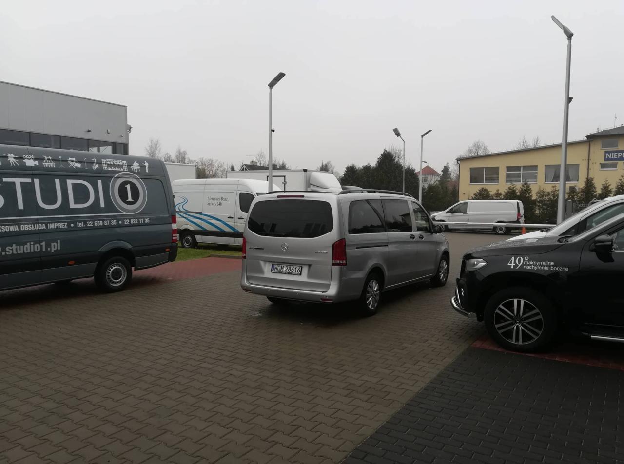 Srebrny Mercedes-Benz Vito zaparkowany na kostce brukowej, widoczny z tyłu, z tablicą rejestracyjną WGM 28616, obok zaparkowane inne busy firmowe, w tle zabudowania.