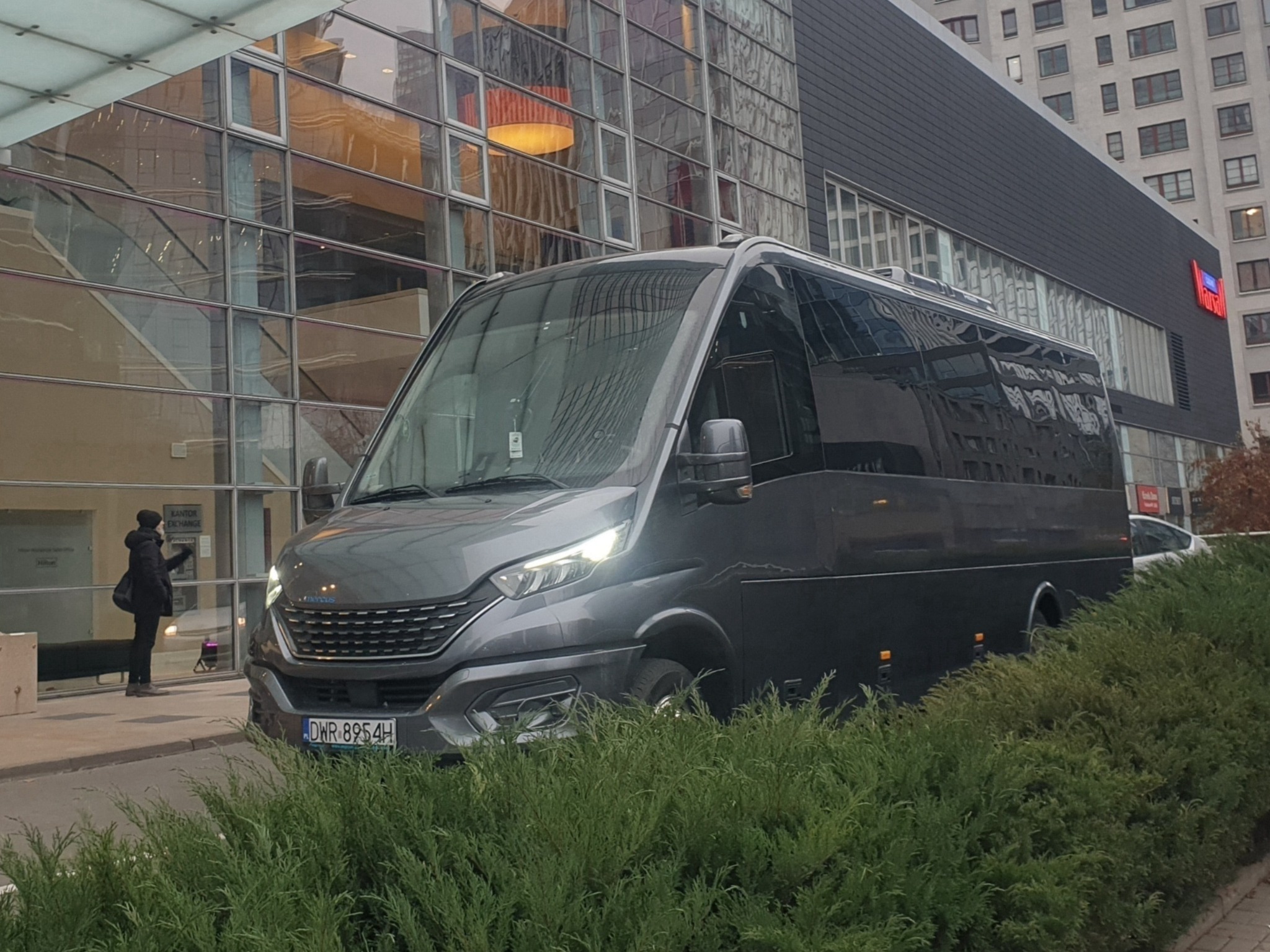 Szary bus Iveco z włączonymi światłami mijania, zaparkowany przed nowoczesnym budynkiem z przeszkloną fasadą, częściowo zasłonięty przez krzewy.