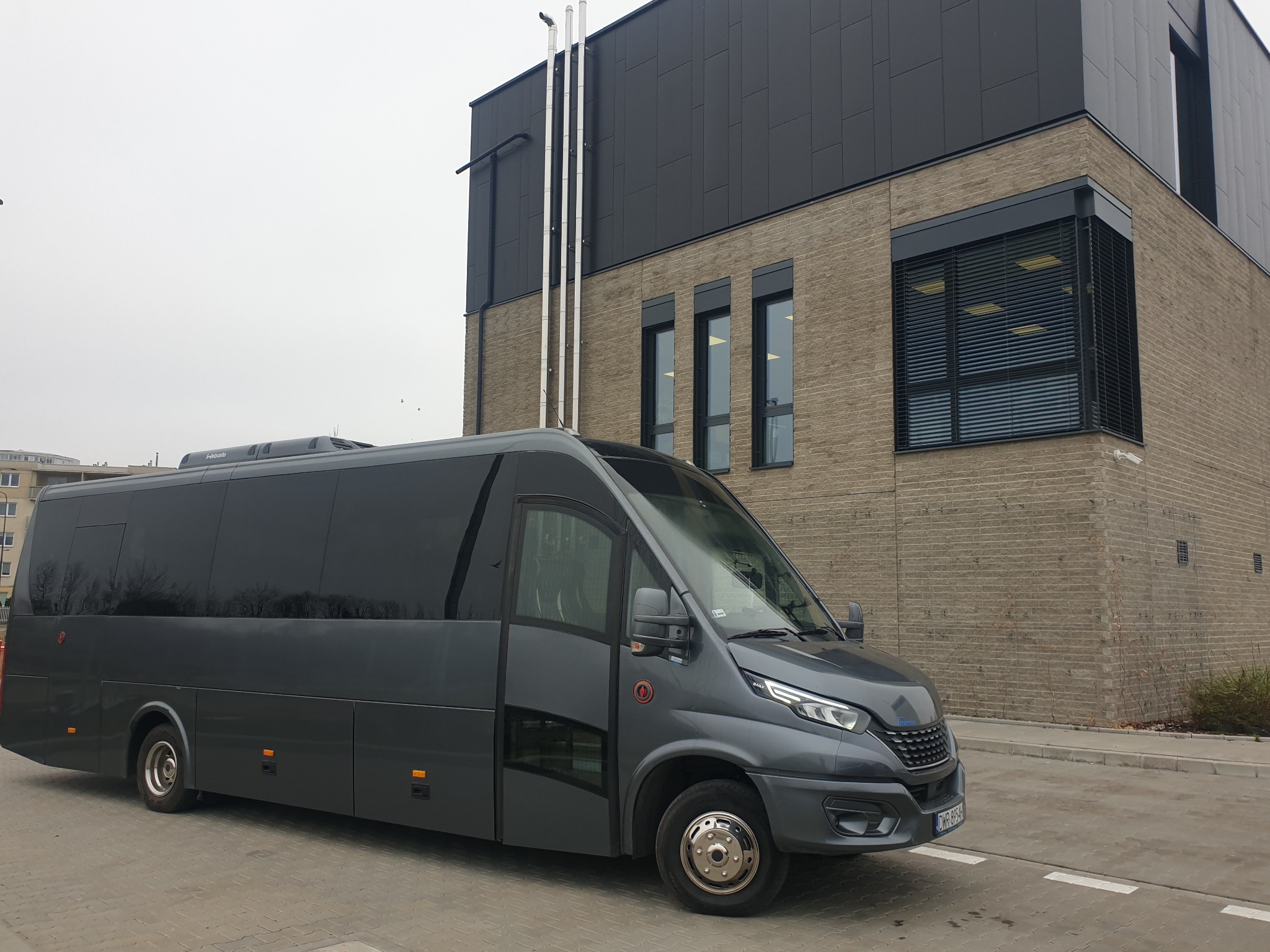 Szary, nowoczesny bus Iveco Daily z przyciemnianymi szybami, zaparkowany na kostce brukowej przed budynkiem z cegły i szarej blachy.