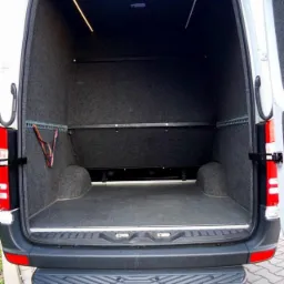 Wnętrze furgonetki wyłożone szarą wykładziną, widok z tyłu na pustą przestrzeń ładunkową z oświetleniem LED i szyną mocującą bagaż.