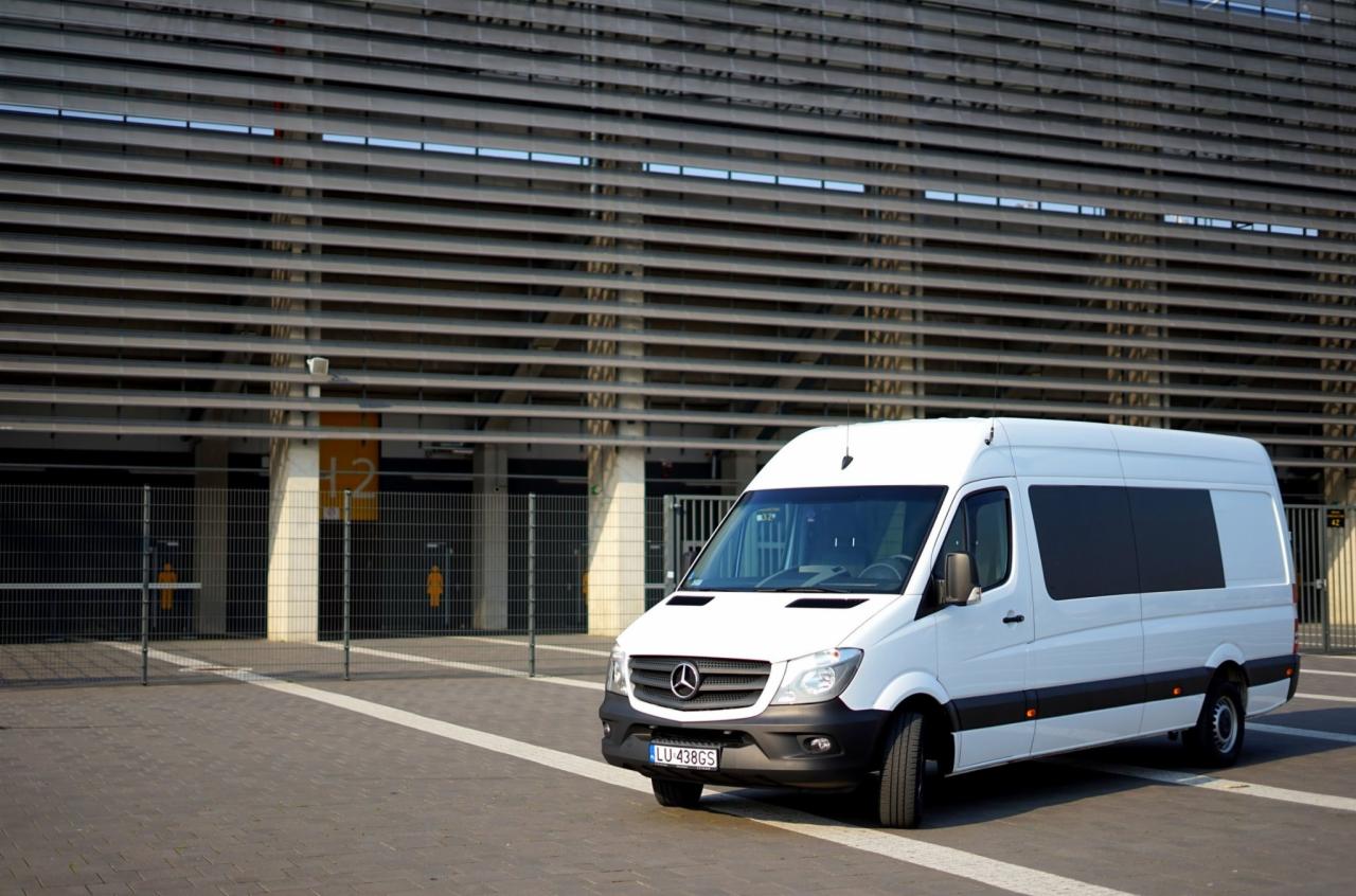 Biały bus Mercedes-Benz Sprinter z przyciemnianymi szybami, zaparkowany na tle nowoczesnej fasady stadionu z metalowymi żaluzjami.