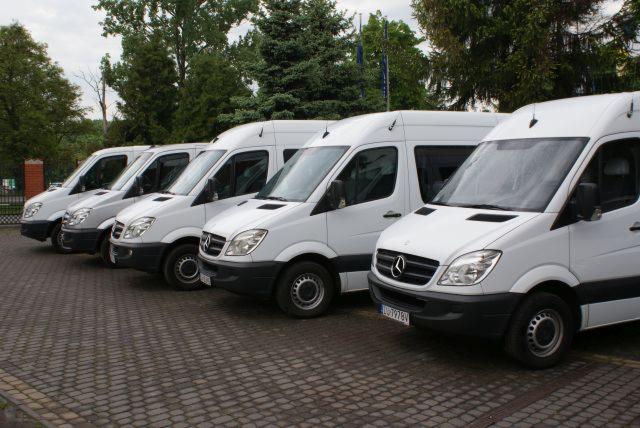 Flota białych busów Mercedes-Benz Sprinter zaparkowana w rzędzie na brukowanym placu, z widocznymi drzewami w tle. Pojazdy wyglądają na zadbane i gotowe do drogi.