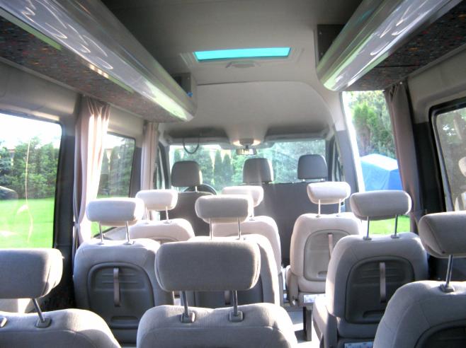 Wnętrze busa pasażerskiego z szarymi fotelami, widok z tyłu na przednie siedzenia i okna z zasłonami. Widoczny świetlik w dachu pojazdu.