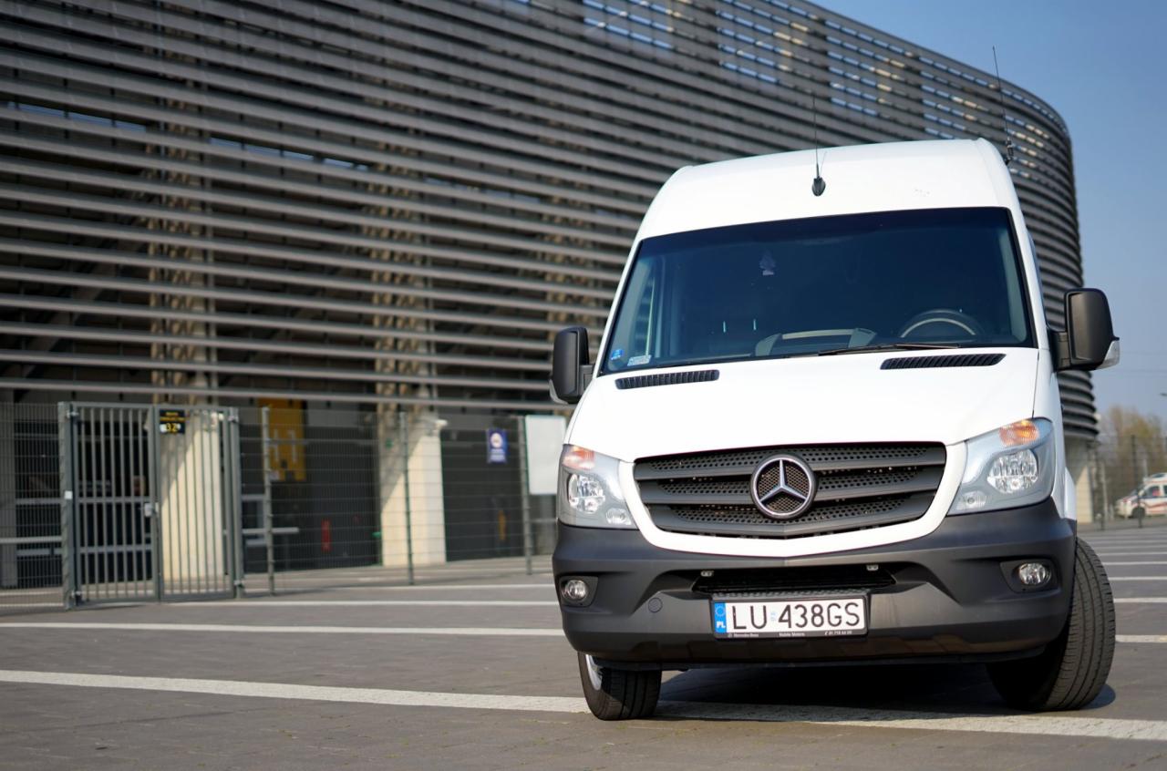 Biały bus marki Mercedes-Benz Sprinter z lubelską rejestracją LU 438GS zaparkowany na placu przed nowoczesnym budynkiem o metalowej elewacji.