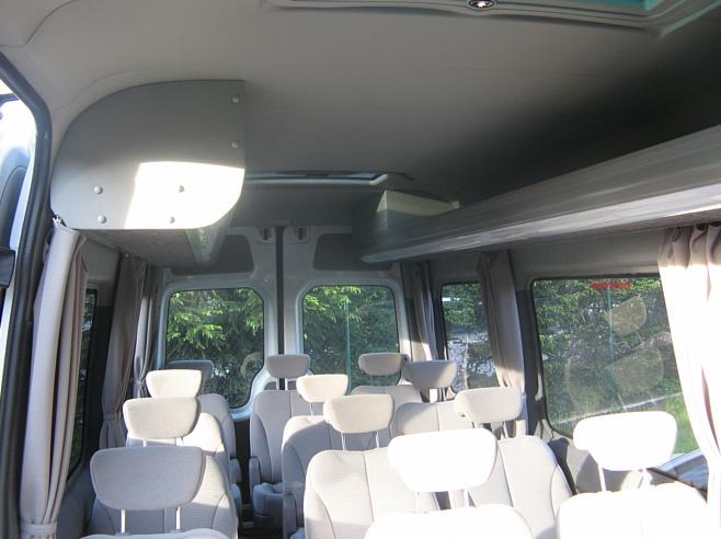 Wnętrze busa pasażerskiego z szarymi siedzeniami i zasłonami w oknach, widok z tyłu pojazdu. Oświetlenie sufitowe i półka nad oknami.