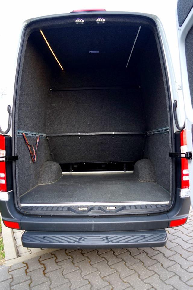Wnętrze furgonetki wyłożone szarą wykładziną, widok z tyłu na pustą przestrzeń ładunkową z oświetleniem LED i szyną mocującą bagaż.