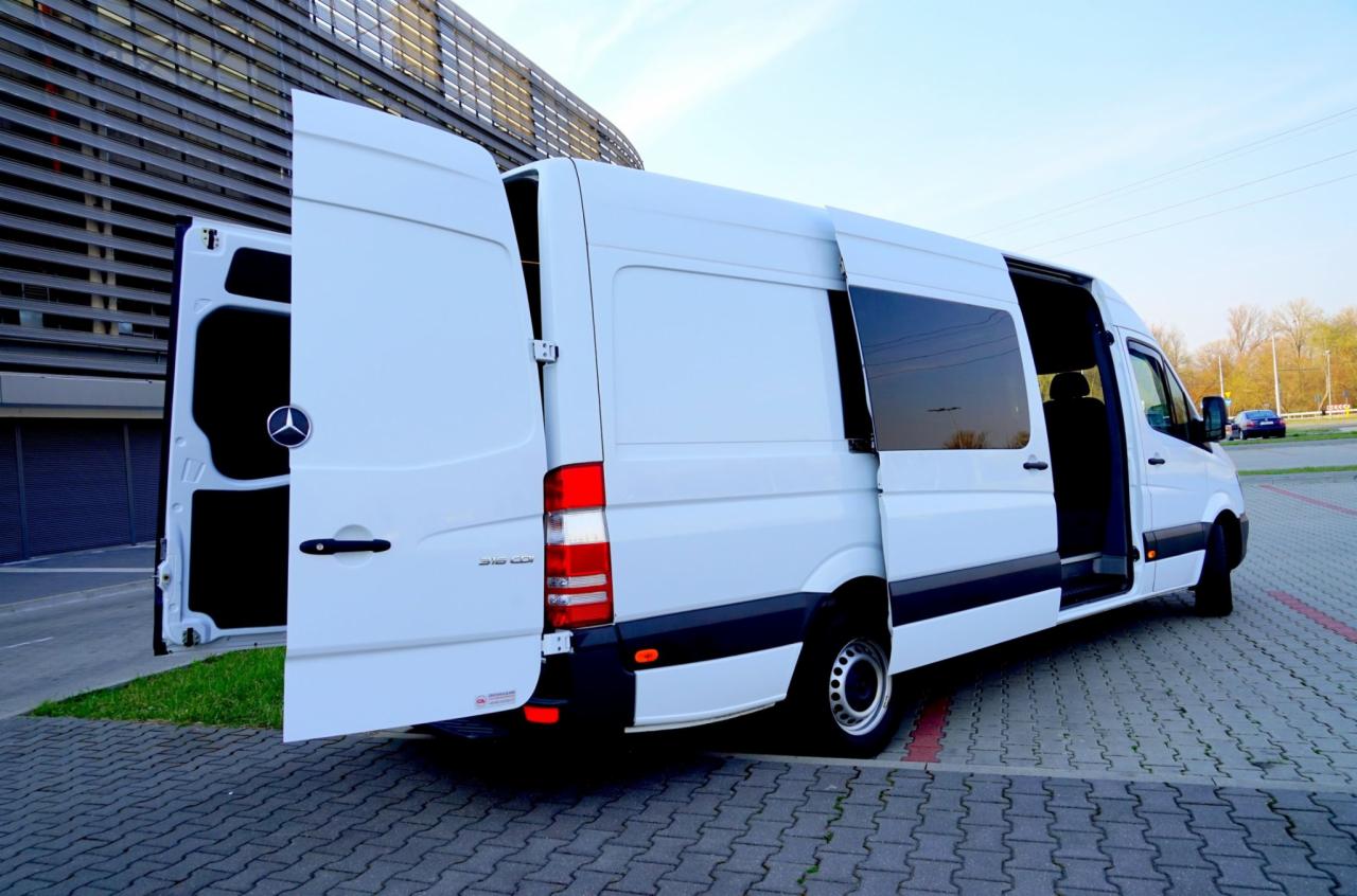 Biały bus marki Mercedes-Benz Sprinter z otwartymi drzwiami bocznymi i tylnymi, zaparkowany na kostce brukowej przed budynkiem.