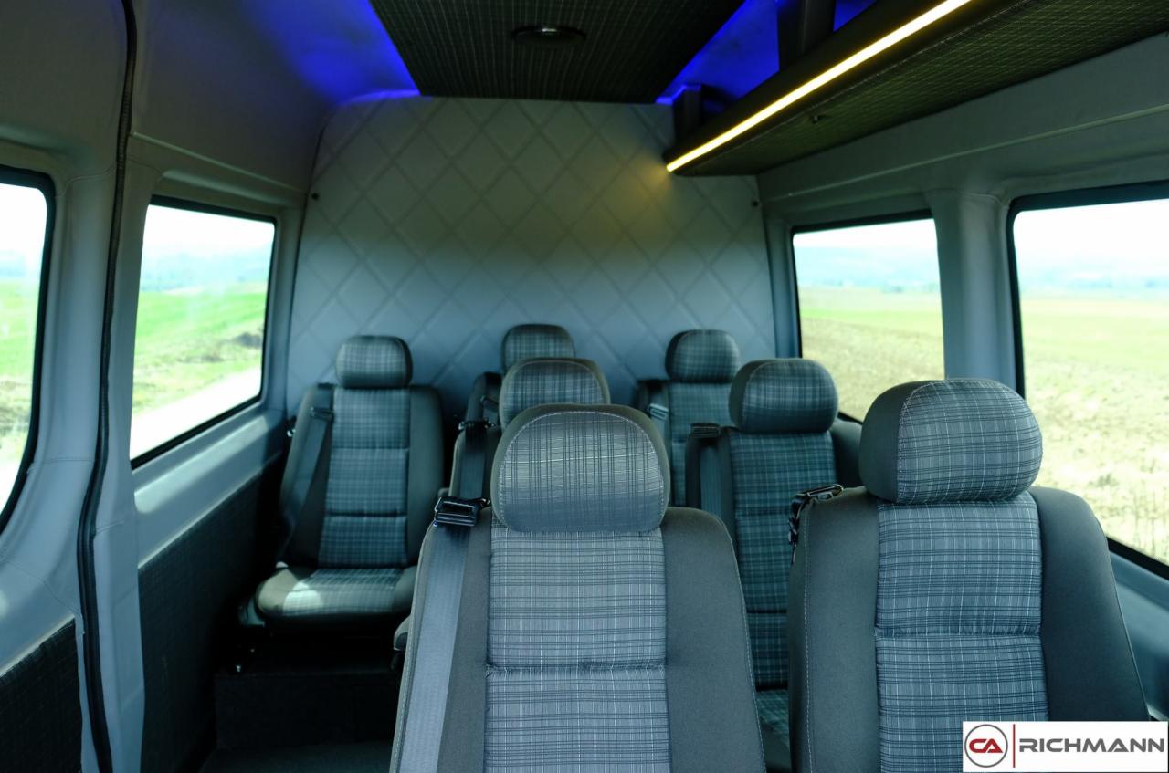 Wnętrze komfortowego busa z szarymi, wzorzystymi fotelami i oświetleniem LED, widok z tyłu pojazdu na zielone pola za oknami.