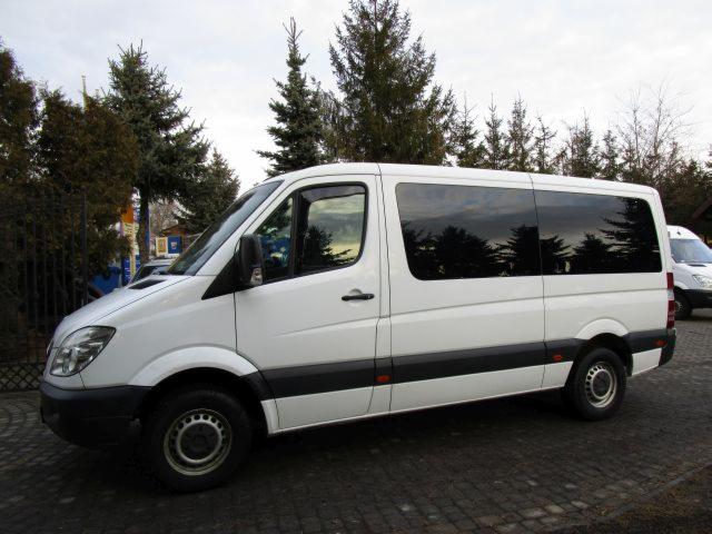 Biały bus Mercedes Sprinter z przyciemnianymi szybami na zewnątrz, zaparkowany na kostce brukowej, otoczony zielenią drzew i krzewów.