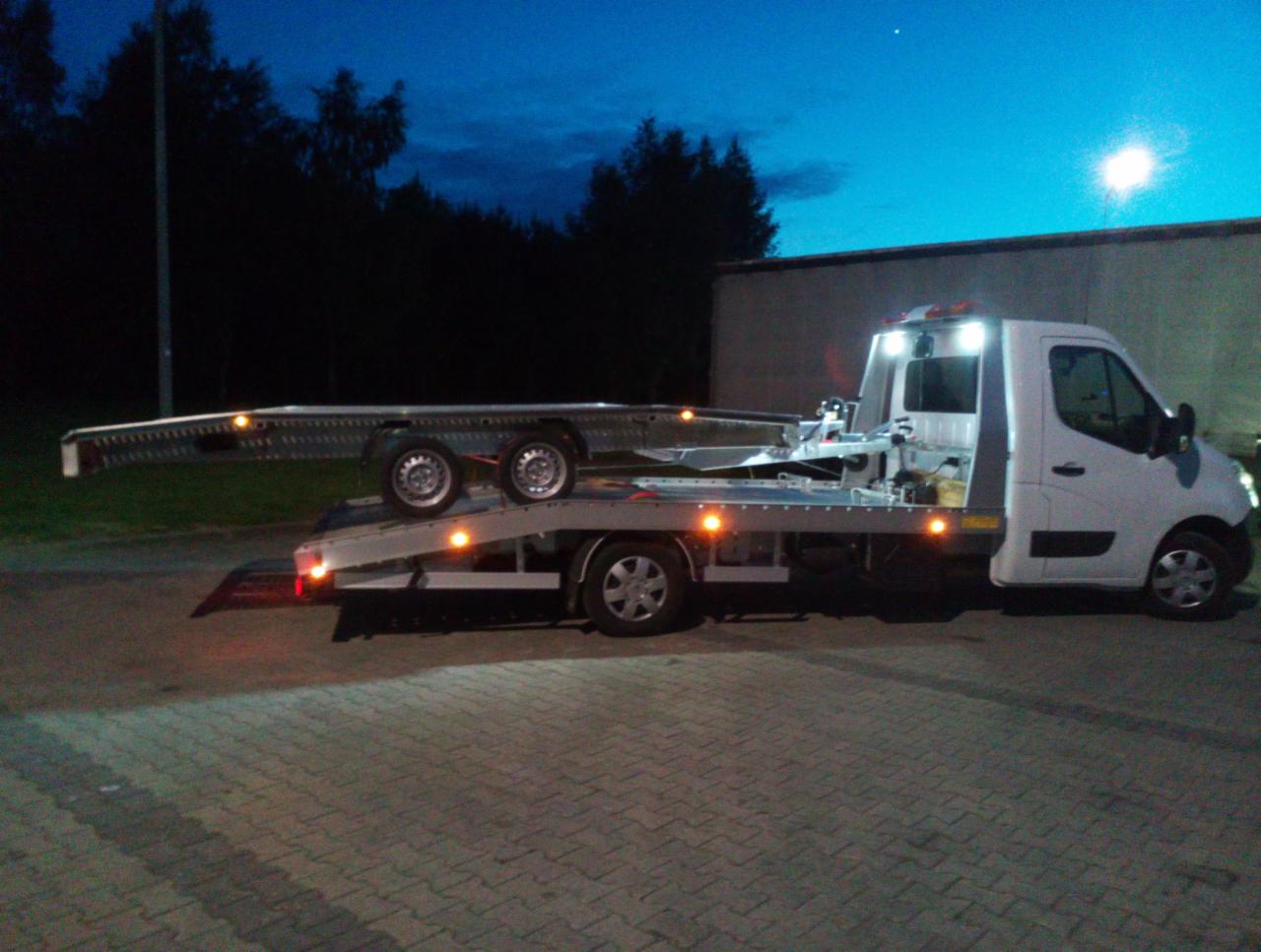 Biała laweta z przyczepą na parkingu o zmierzchu, oświetlona światłami. Widoczne drzewa w tle i budynek. Przyczepa pusta, gotowa do załadunku.