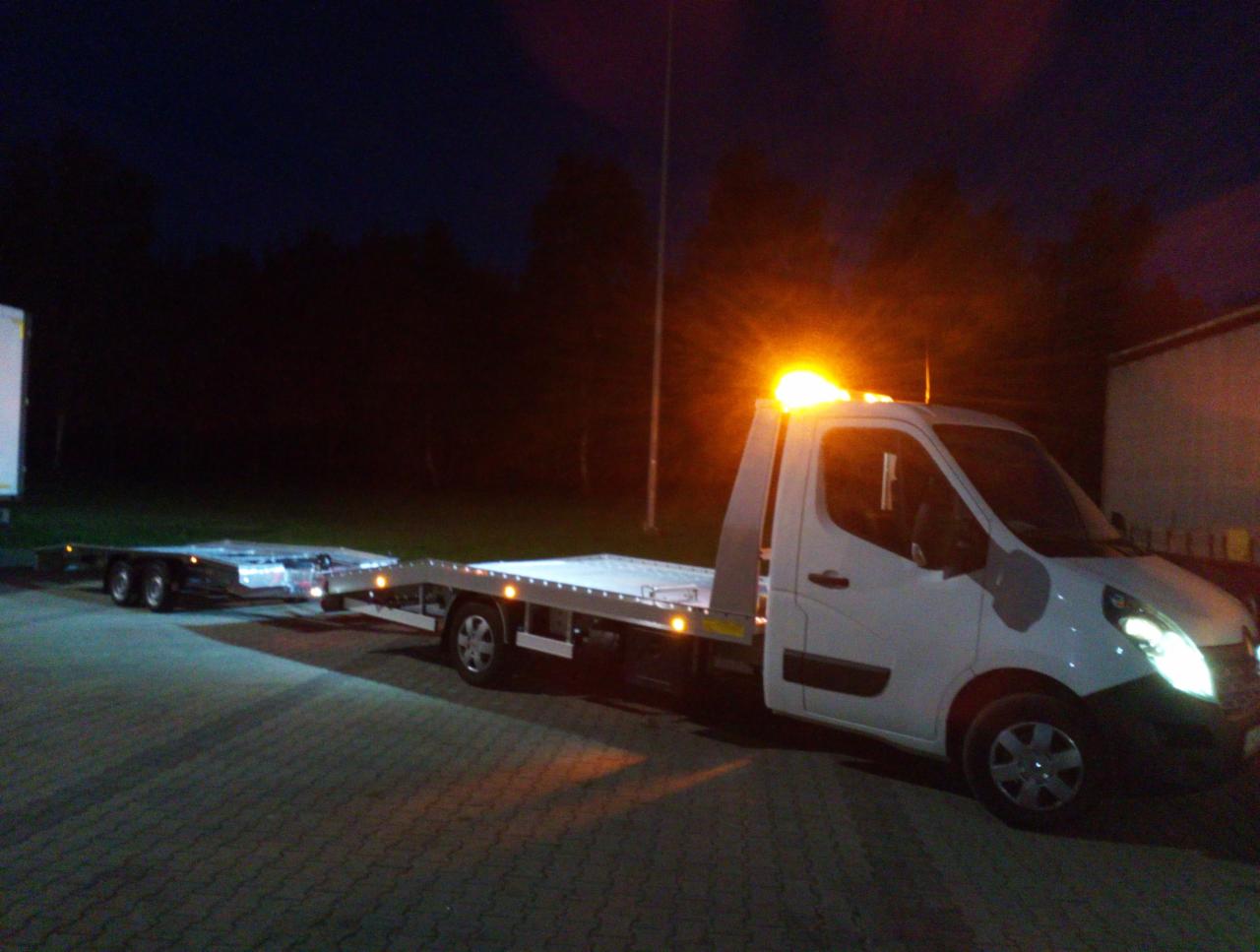 Biała laweta z włączonymi światłami awaryjnymi, zaparkowana na placu w nocy. Widoczna przyczepa lawety i oświetlenie.