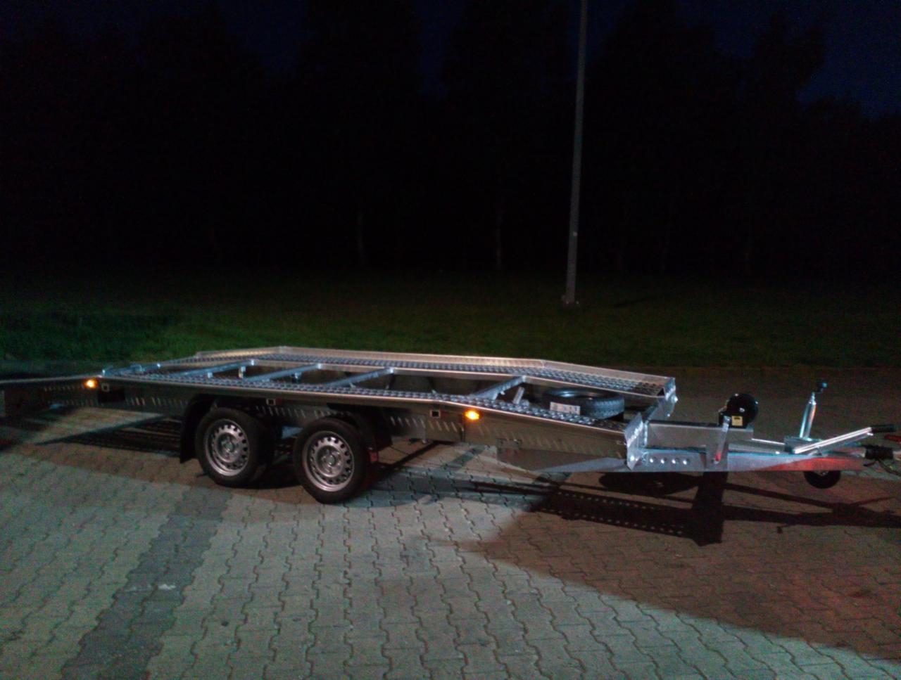 Nowa, aluminiowa laweta samochodowa z oświetleniem pozycyjnym, zaparkowana na kostce brukowej nocą, z widocznymi detalami konstrukcyjnymi i kołem zapasowym.