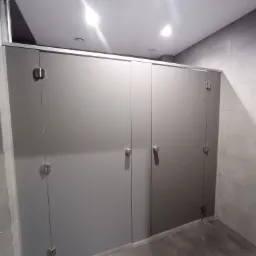 Publiczna toaleta z kabinami o gładkich, szarych drzwiach z metalowymi zawiasami i uchwytami, widoczne płytki podłogowe i ścienne.