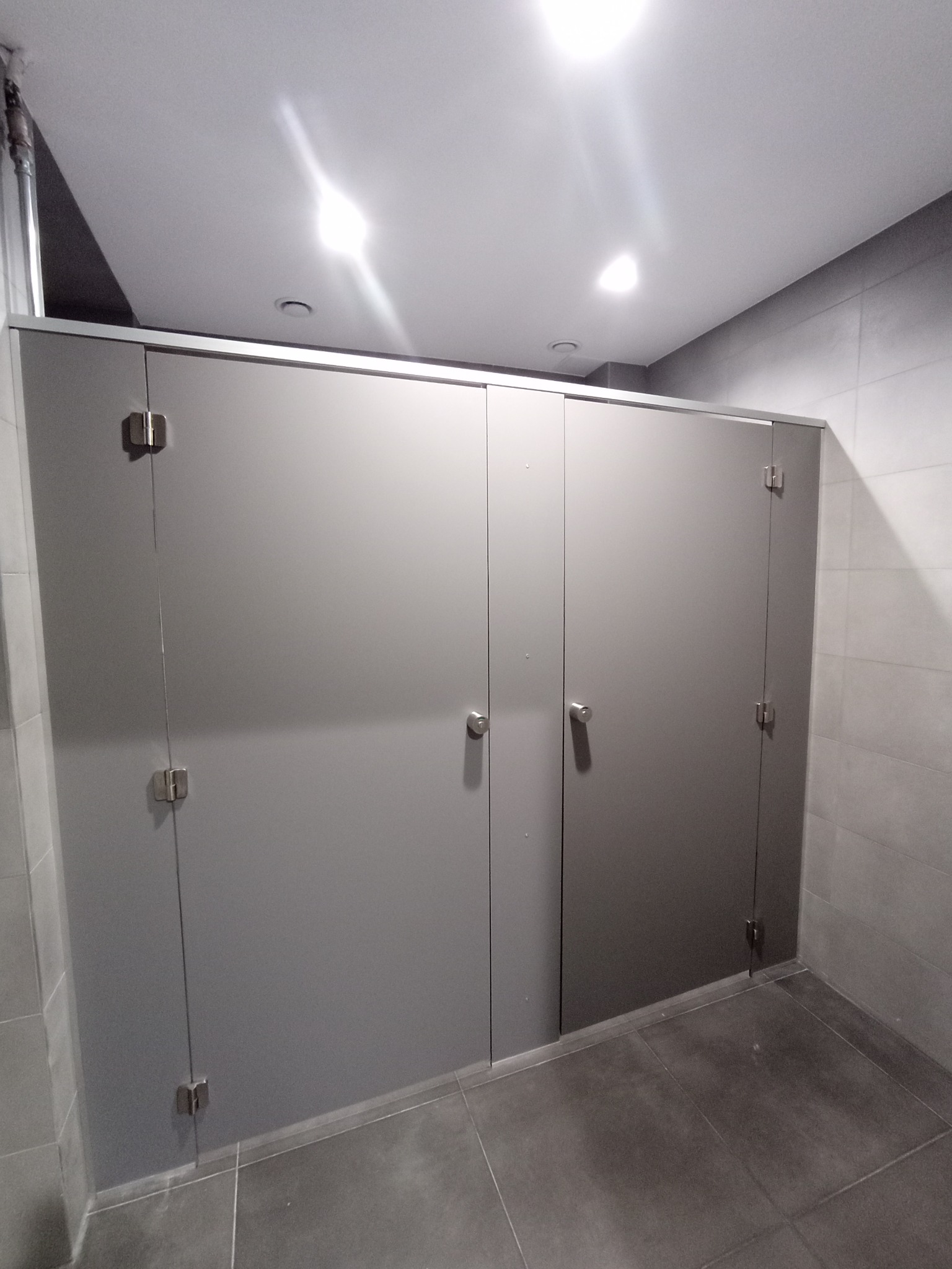 Publiczna toaleta z kabinami o gładkich, szarych drzwiach z metalowymi zawiasami i uchwytami, widoczne płytki podłogowe i ścienne.
