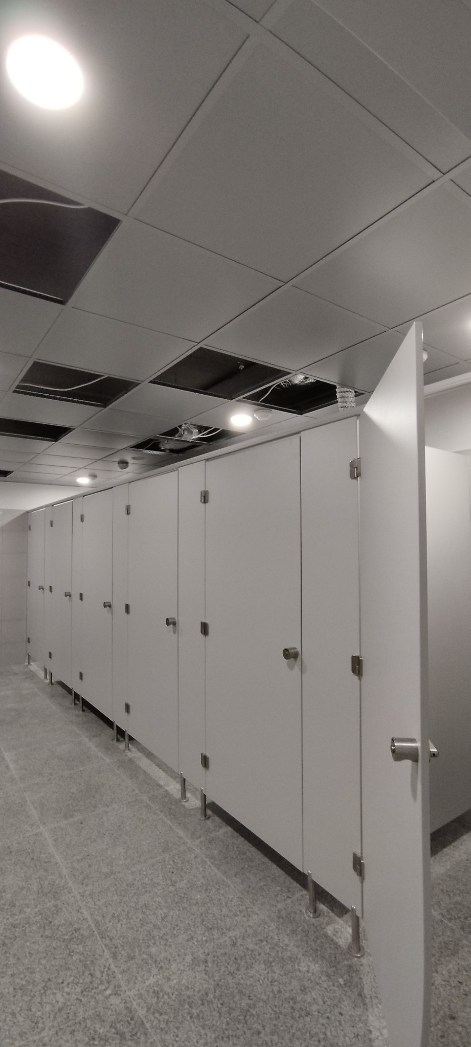 Rząd kabin toaletowych z białymi drzwiami i metalowymi uchwytami, widoczne elementy instalacji podwieszanego sufitu.