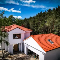 Nowoczesny, dwukondygnacyjny dom z czerwonym dachem z dachówki ceramicznej i oknem dachowym, otoczony zielenią drzew, widziany z lotu ptaka w słoneczny dzień.