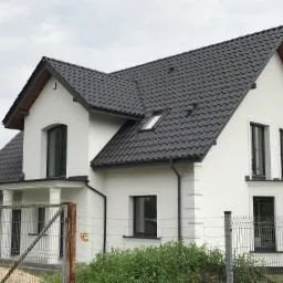 Nowoczesny dom jednorodzinny z ciemną dachówką ceramiczną, białymi ścianami i czarnymi ramami okiennymi, widziany zza ogrodzenia z siatki.