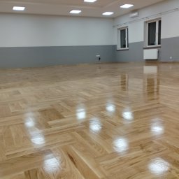 Sebastian Kazior - Jasna, parkietowa podłoga ułożona we wzór jodełki w przestronnym, odnowionym wnętrzu z szarą lamperią i białymi ścianami. Widoczne okna i grzejniki.