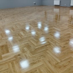 Sebastian Kazior - Parkiet ułożony w jodełkę, po renowacji. Jasne drewno z widocznym usłojeniem, błyszcząca powierzchnia odbija światło. Szara ściana i białe grzejniki w tle.