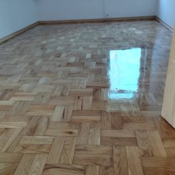 Sebastian Kazior - Parkiet ułożony w jodełkę, świeżo polakierowany, z odbiciem okna. Widoczna część drzwi po prawej stronie kadru. Jasne drewno i ściany.