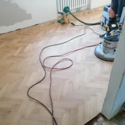 Parkiet ułożony w jodełkę, w trakcie prac wykończeniowych, widoczna maszyna do szlifowania i odkurzacz przemysłowy, kabel zasilający na podłodze.