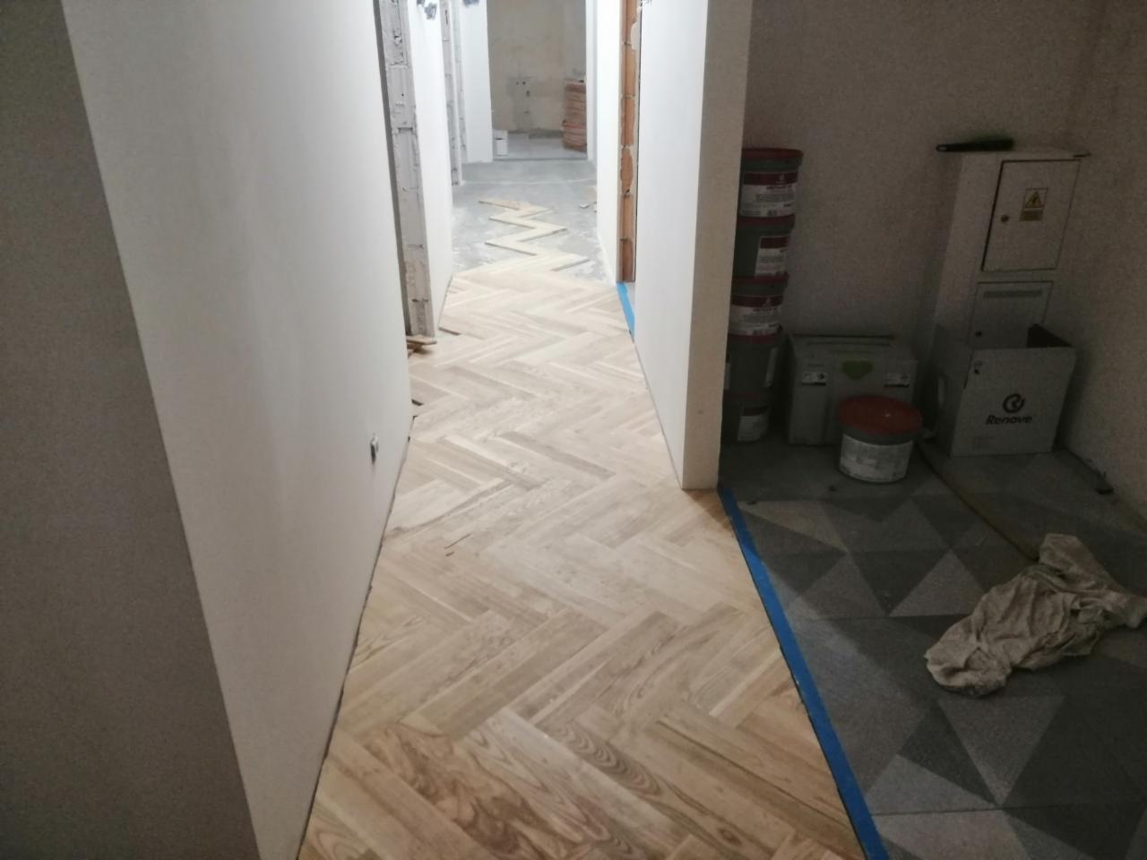 Wnętrze w trakcie remontu: ułożona drewniana podłoga w jodełkę w korytarzu, widoczne puszki z farbą i narzędzia malarskie, zabezpieczona folią podłoga w sąsiednim pomieszczeniu.