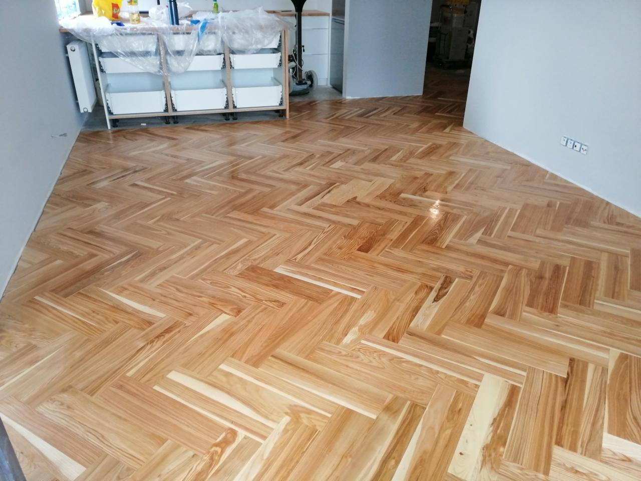 Nowo ułożona podłoga z parkietu w jodełkę, jasne drewno z wyraźnym usłojeniem, widoczne gniazdka elektryczne na ścianie, w tle białe meble.