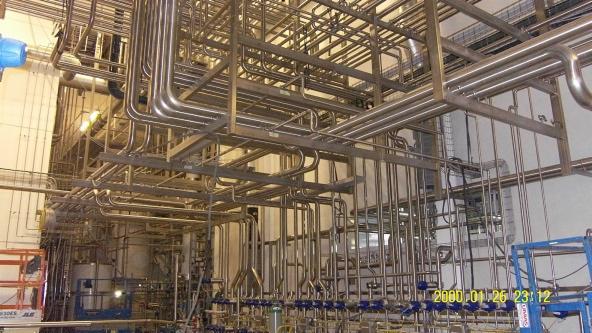 Złożona instalacja rurociągów ze stali nierdzewnej w hali przemysłowej, z licznymi zaworami i elementami łączeniowymi, widoczne podnośniki koszowe.
